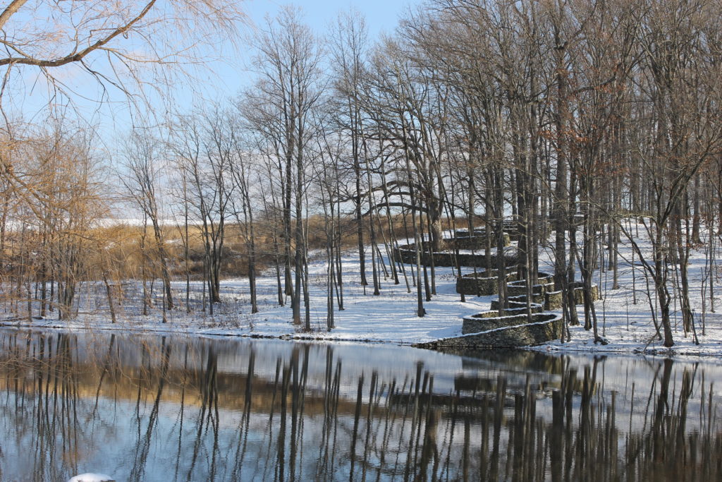 Snowy landscape with bare trees and a curving stone wall reflected in a pond.