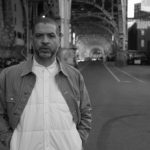 Portrait of Jason Moran standing on a city street under a bridge overpass.
