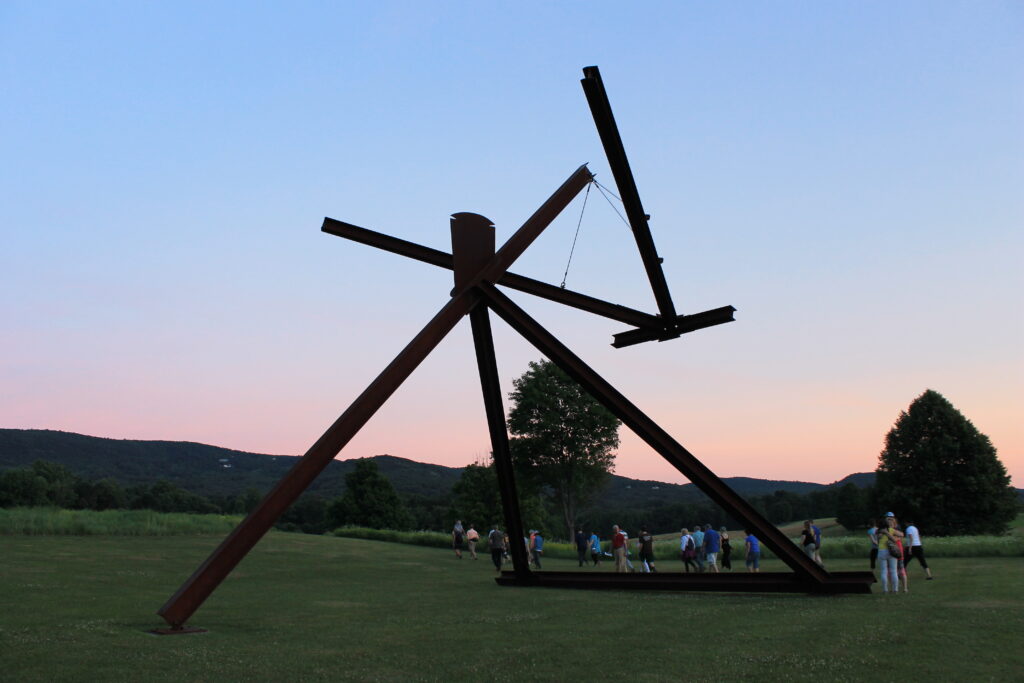 A group of people walk past a large-scale sculpture made of steel beams at sunset
