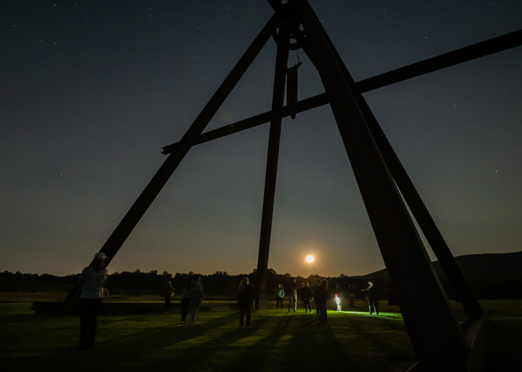 in a night time setting with the moon rising over the horizon, a group of people stand inside a massive, hollow, pyramidal metal sculpture while listening to a tour leader speak about the work