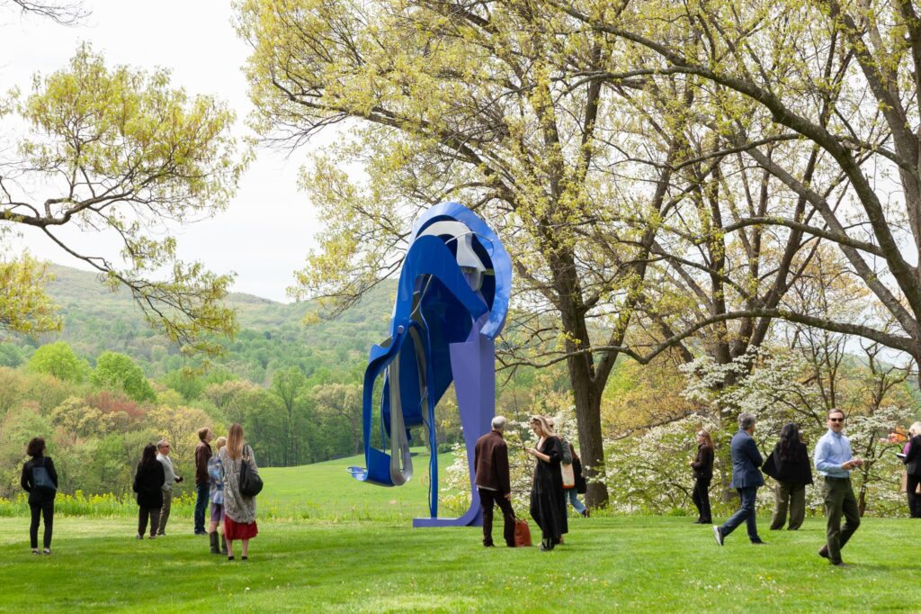 Visitors walk around a tall steel sculpture standing in a grassy lawn, with blooming trees above