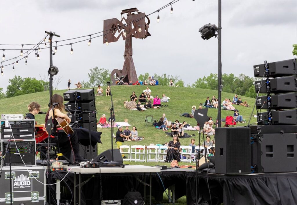 an outdoor music stage with people sitting on the grass among a large-scale sculpture