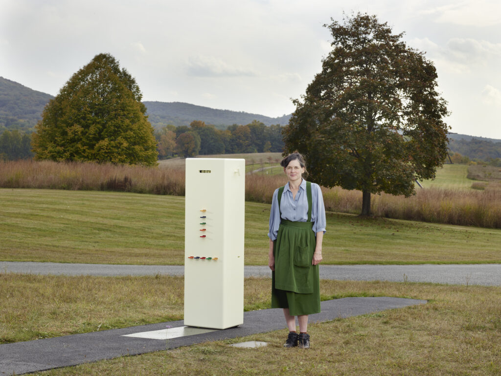 Artist Taryn Simon standing near her sculpture, Kleroterion, a cubic white structure with multiple slots and chips.