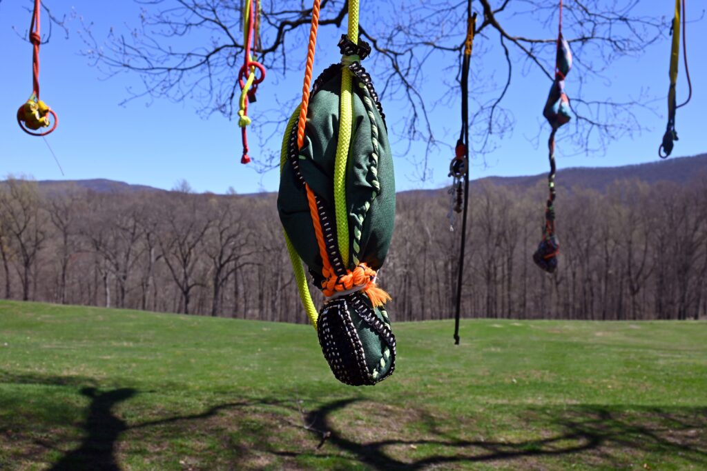 a banana shaped hanging sculpture made of ropes and textiles, suspended from a tree on a grassy hill.