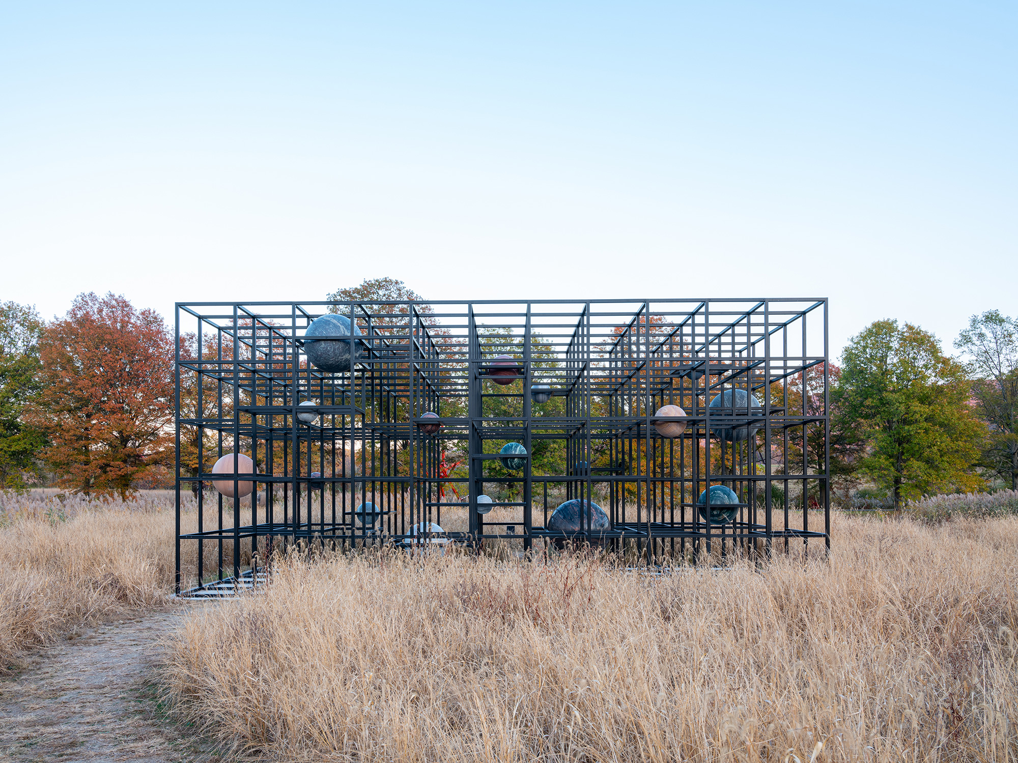 In a field of native grass sits a sculpture made of a grid of black steel, with round natural stones suspended inside.