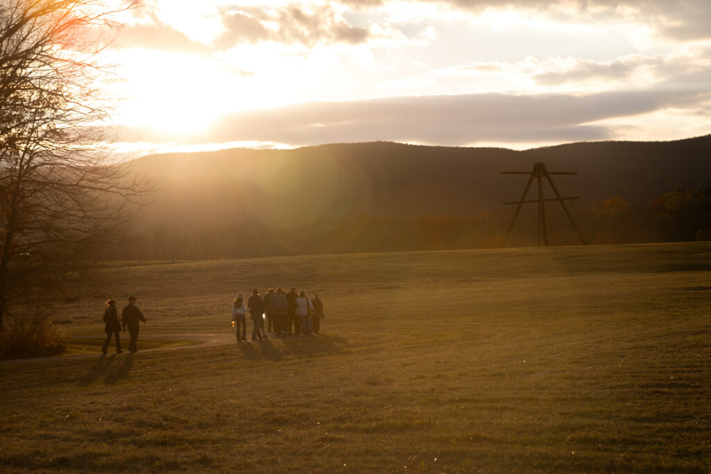 A tour group walks through a sunset-drenched meadow, bordered by a mountain range in the distance.