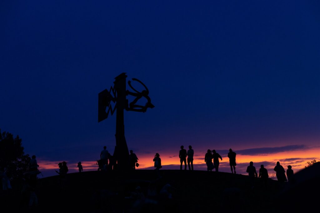 At nightfall with an orange sunset glow on the horizon, silhouettes of people stand atop a round hill among a tall sculpture.