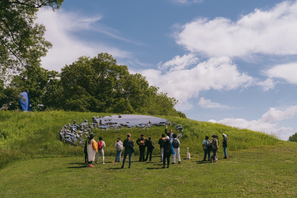 A group of visitors attend a tour. They are looking up a hill at a stainless steel structure arranged in a ring, with over 100 different pieces scattered like an archipelago, each covered with a mirror-reflective surface such that the sky above is visible in each one. Lush grasses rise in the cracks and gaps to give the effect of a ruin, a mysterious object fallen to the earth, constantly reflecting the light and motion of the atmosphere.
