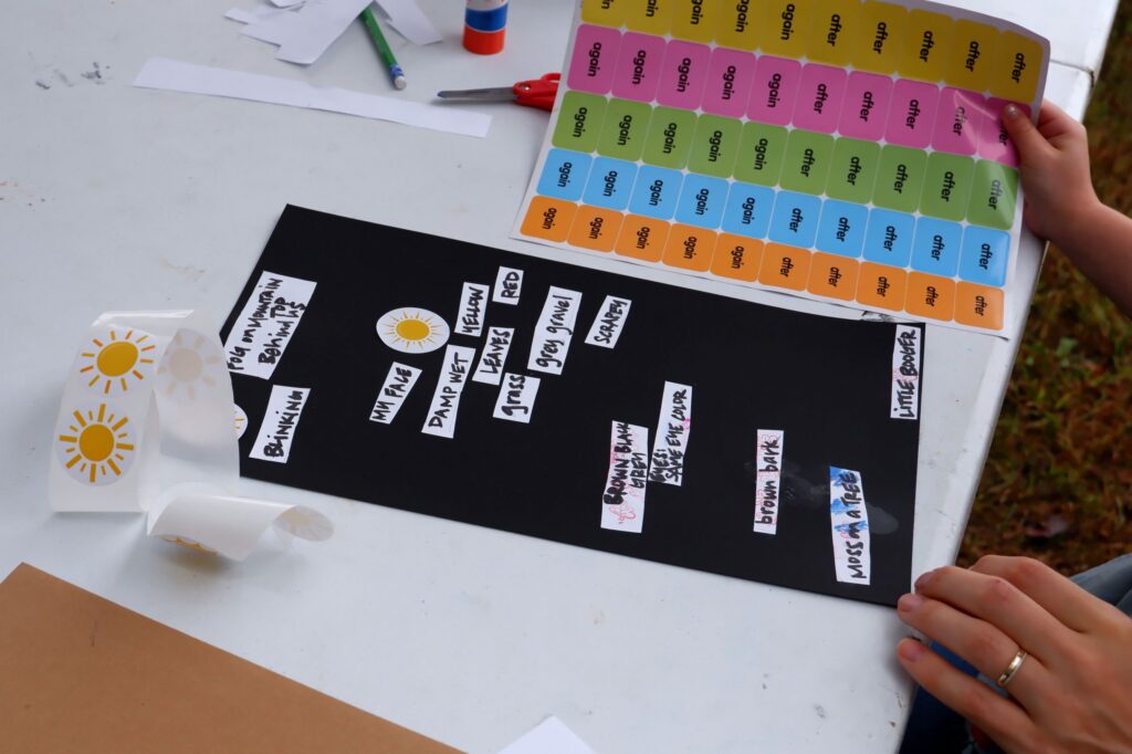 Hands making a poem poster on black cardstock using cut out words on white paper and stickers.