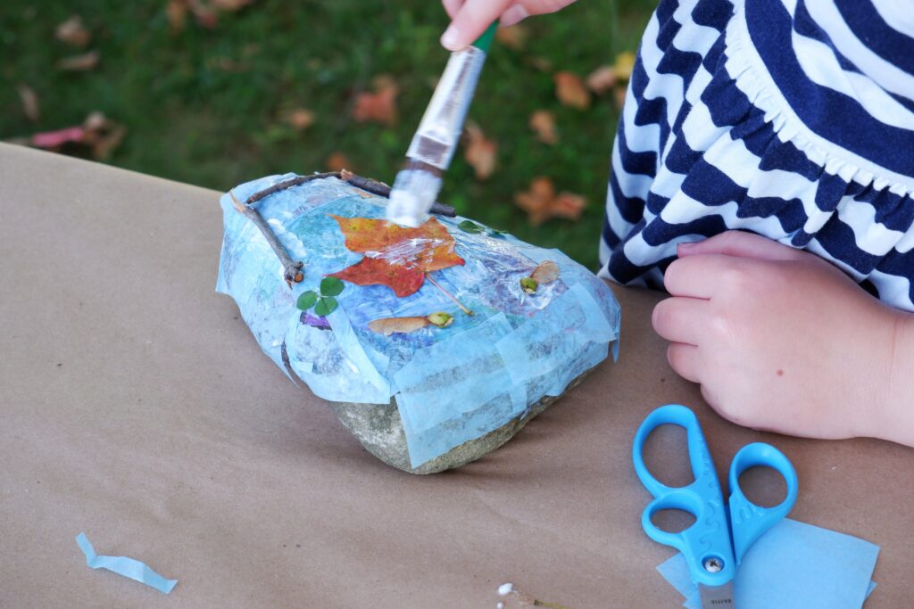 Hands of a young child using a paint brush to paint a leaf on a rock. The rock sits on a table covered in brown paper.