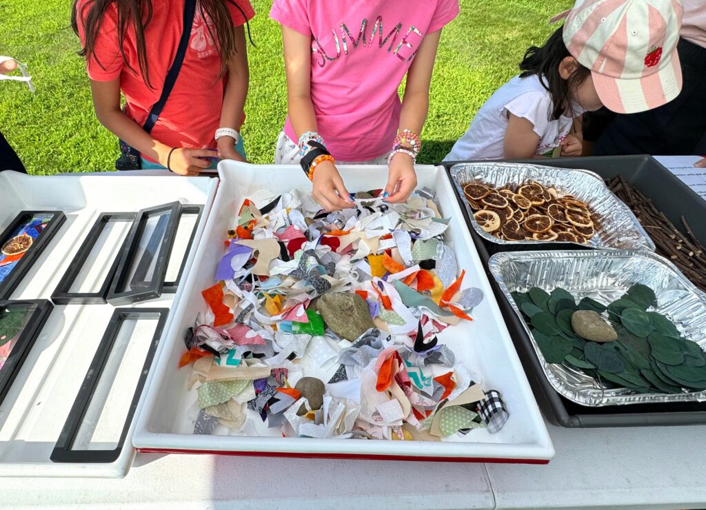 Small children stand in front of a table that has trays of bright colorful fabric and paper pieces.