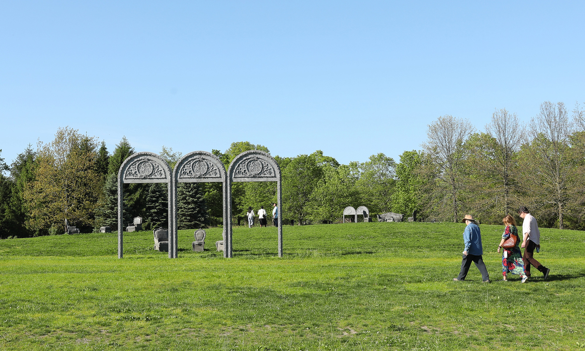 Three decorative arches stand in a grassy meadow, surrounded by grey concrete chairs and couches. People walk through the meadow
