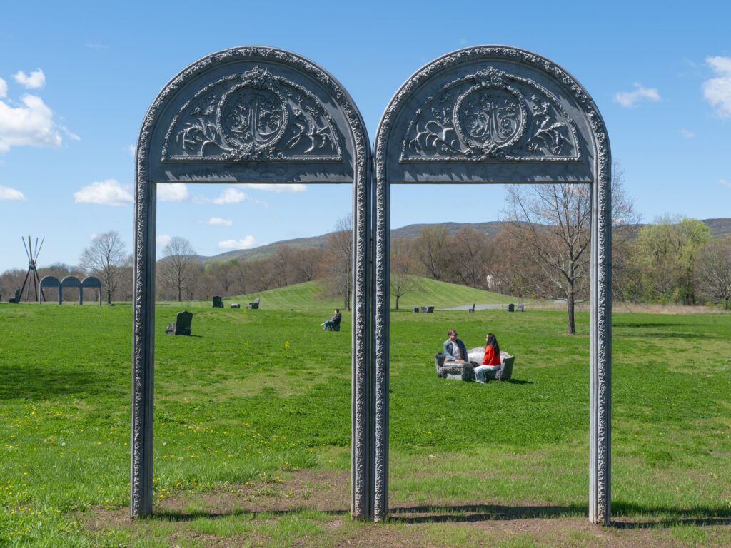 Two decorative arches stand in a grassy meadow, surrounded by grey concrete chairs and couches. People are sitting on one of the couches, while a goose wanders nearby.