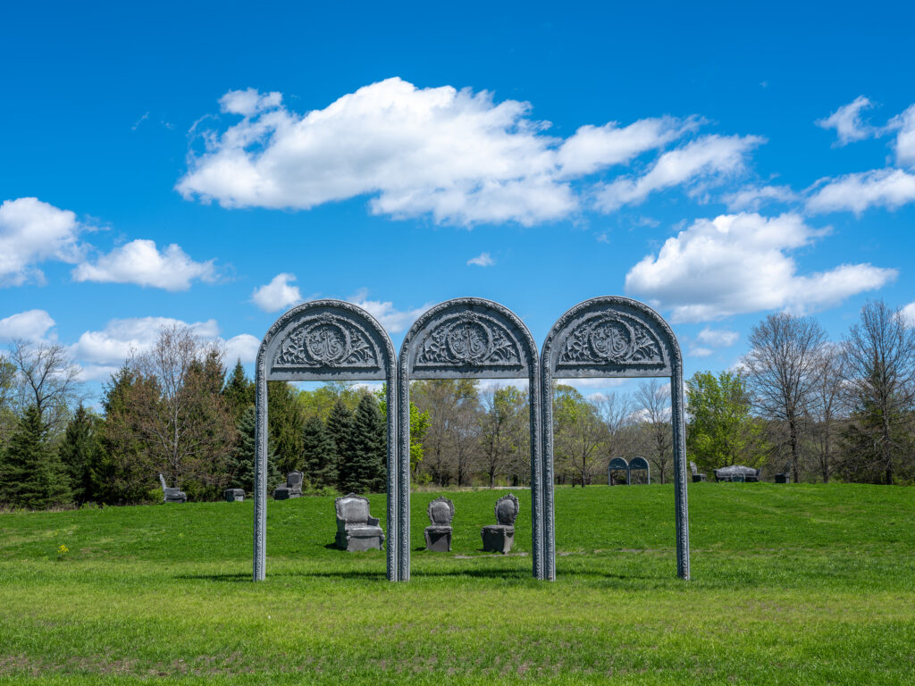 Three decorative arches stand in a grassy meadow, surrounded by grey concrete chairs and couches.