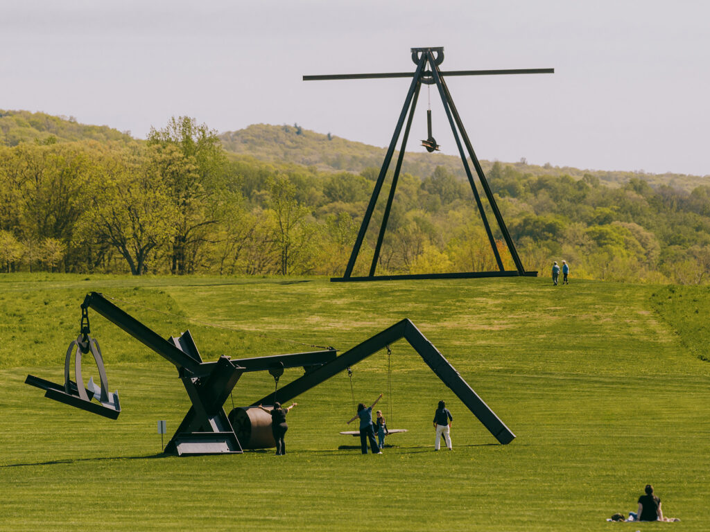 A family plays on the swing attached to a steel sculpture. In the bottom right corner, a woman sits on the grass. In the distance a couple walks near a very large steel sculpture.