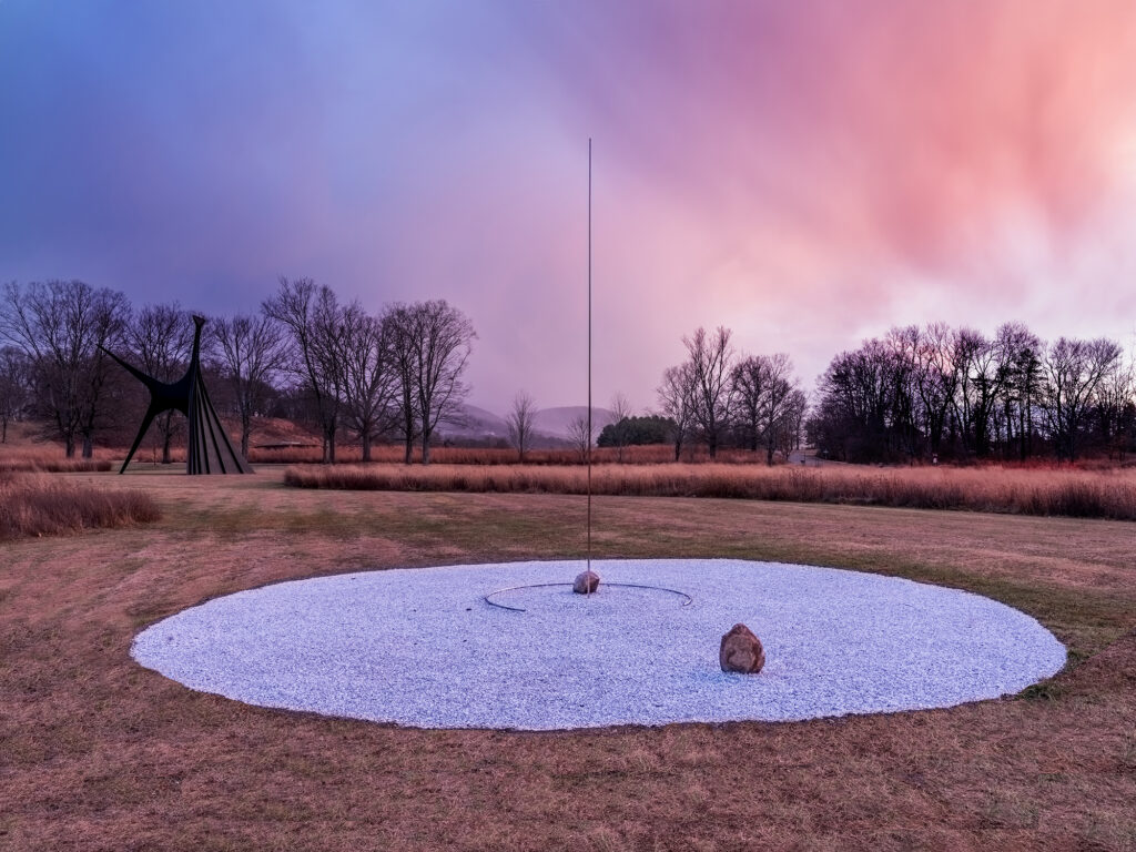 In a field of browning grass, sits a large flat circle of small white stones. At the center is a large rock, with a vertical steel pole. Another large rock sits off center in the circle.