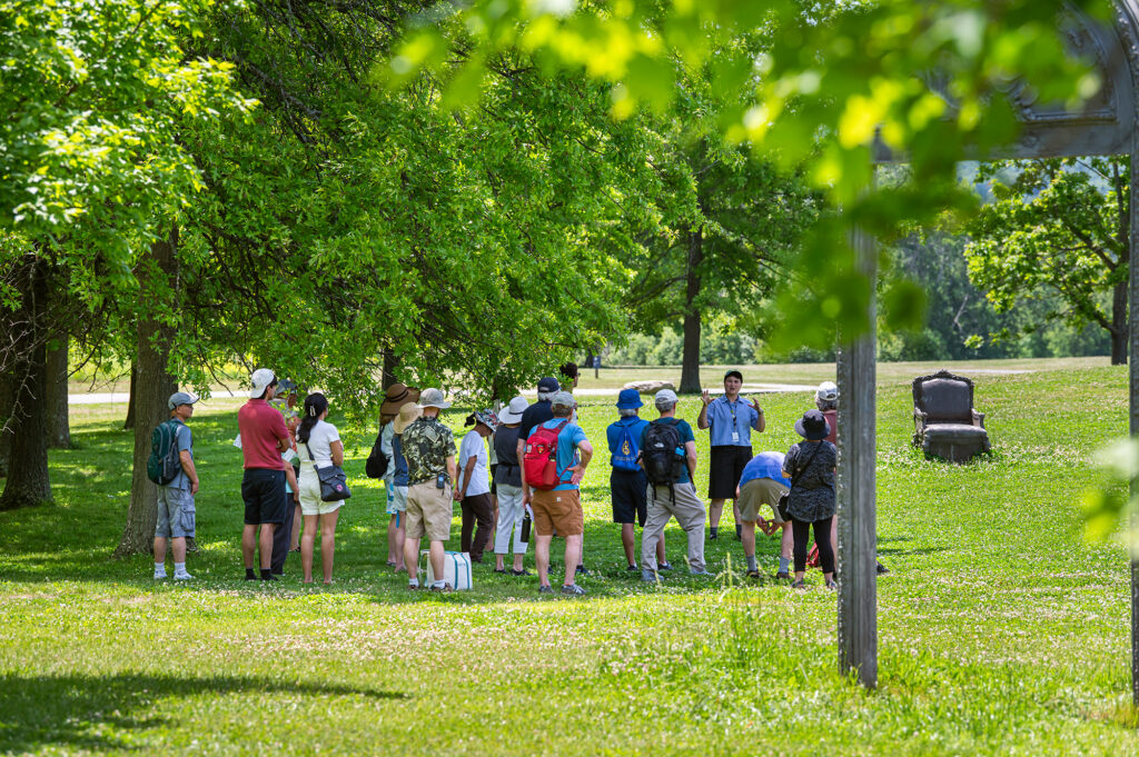 A group of people are on a tour in a grassy field. The tour leader is gesturing with their hands. A concrete chair rests in the filed, with part of a concrete arch in the foreground.