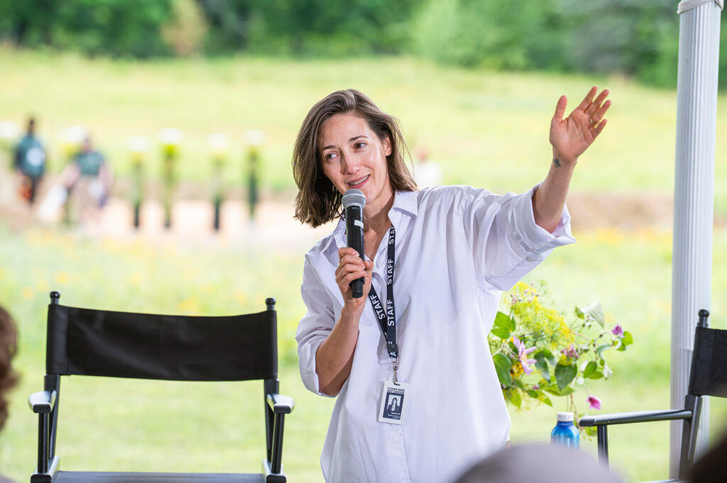 A woman speaks into a microphone. They are wearing a white shirt, with a black lanyard that says staff.