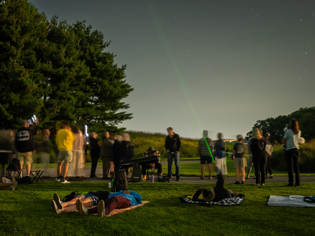 A group of people look at the night sky. Two people in the foreground lay on a blanket looking up. A person in the middle looks through a telescope. 