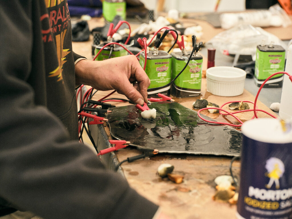 A close up on a hand wiping a substance over a metal sheet. Large batteries sit in the background.
