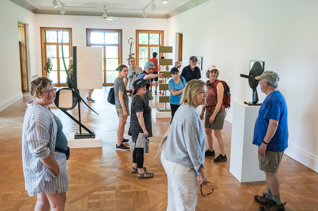 In an indoor gallery, a group of stand around several steel sculptures that stretch from the ground or rest on pedestals